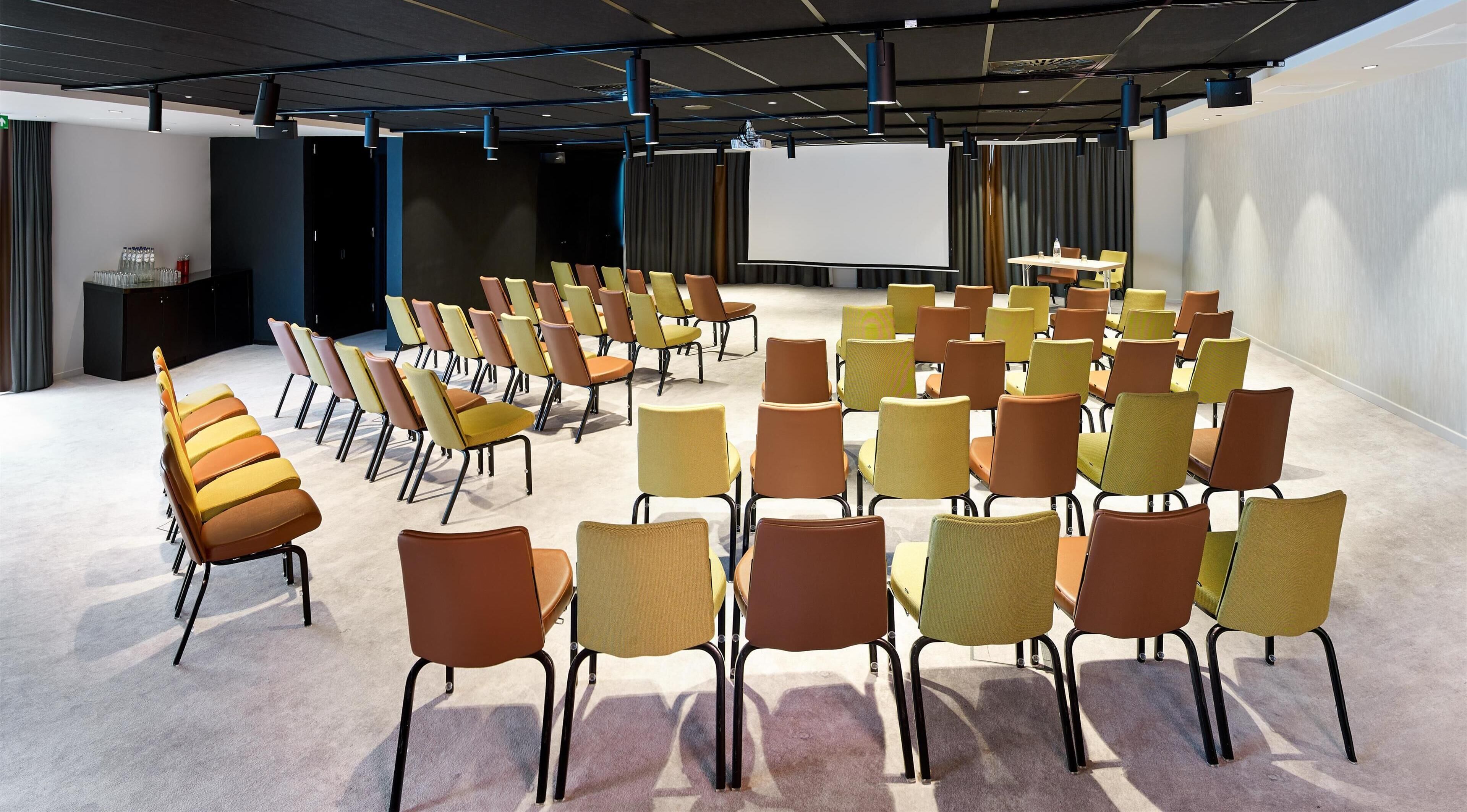 A conference room with rows of chairs, a projector screen, and a table with a bottle.