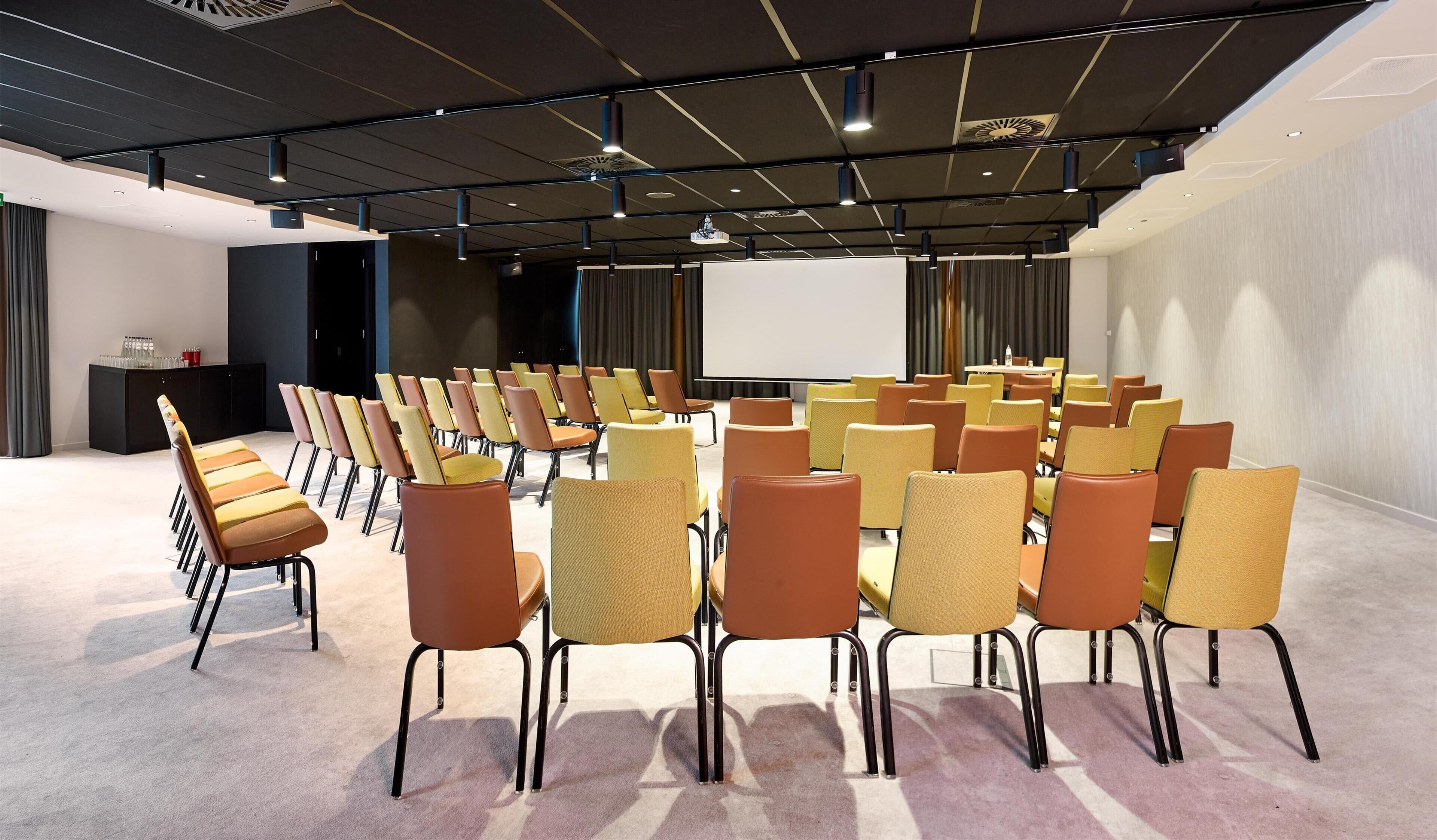 Un salón de conferencias con filas de sillas, una pantalla para proyector y un atril.