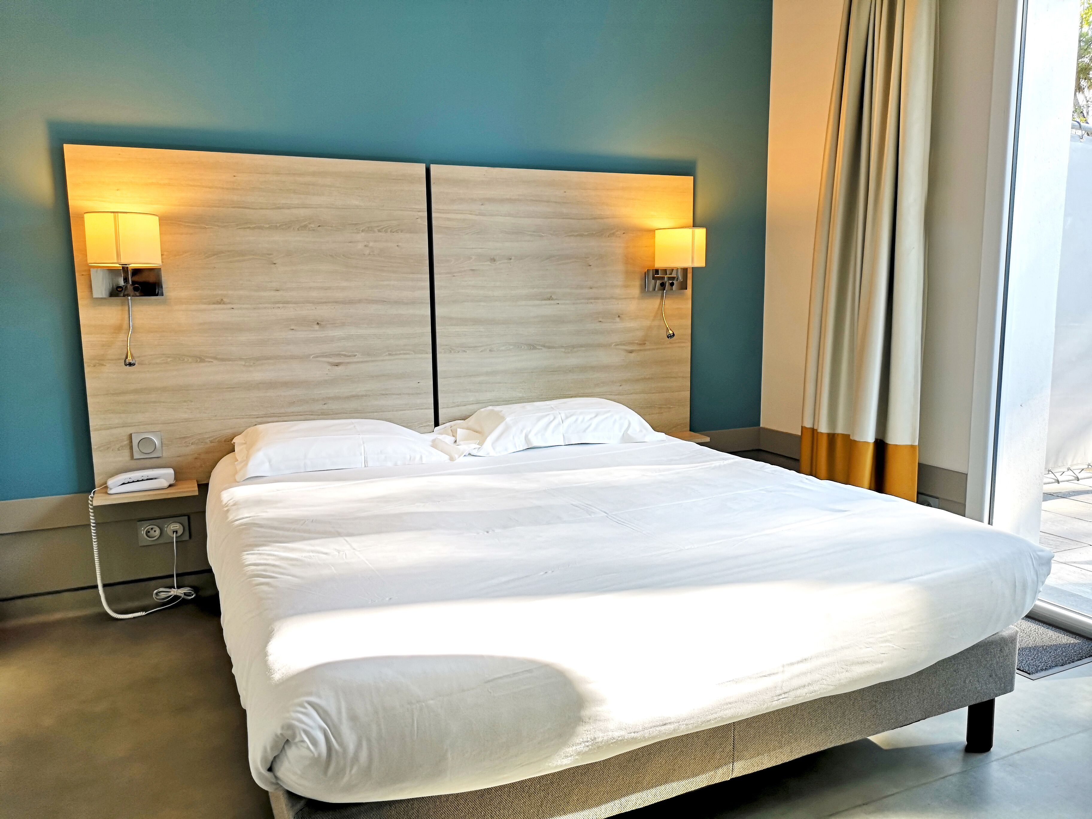 A hotel room with a large bed, two bedside lamps, a bedside phone, and a window with curtains.