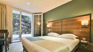 Een hotelkamer met een groot bed, een bureau en uitzicht op bomen door de glazen deur.