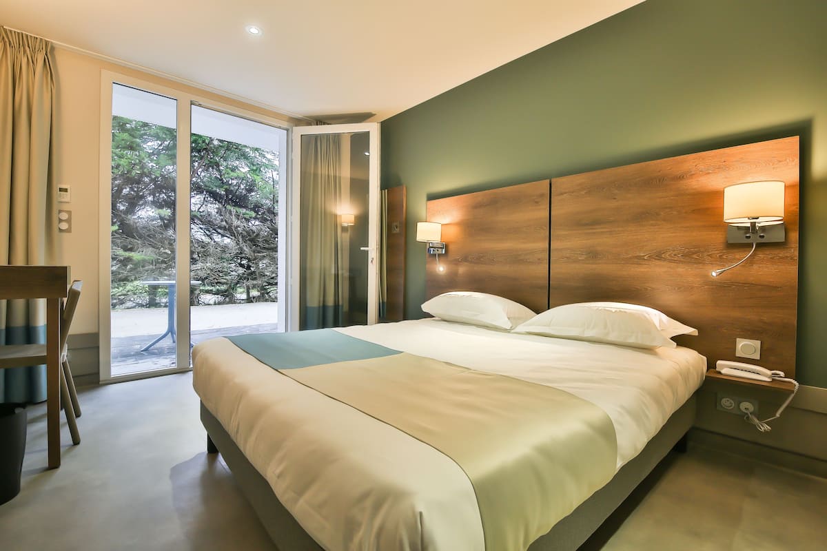 Ein Hotelzimmer mit einem großen Bett, einem Schreibtisch und Blick auf Bäume durch die Glastür.