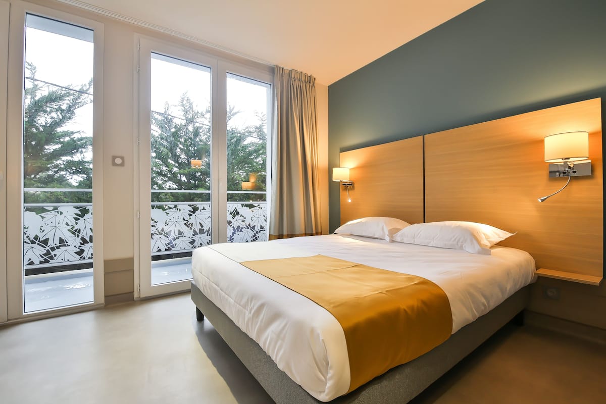 Ein modernes Hotelzimmer mit einem großen Bett, einem Holz-Kopfteil und Blick durch eine Schiebetür auf einen Außenbalkon.