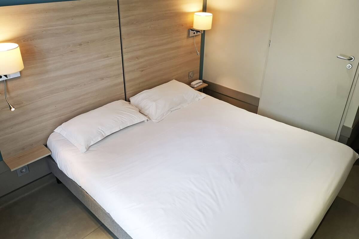 Ein ordentlich bezogenes Bett mit weißen Laken, zwei Nachttischlampen und einem hölzernen Kopfteil.