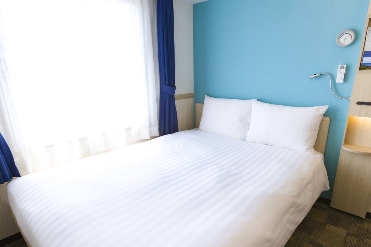 A hotel room with a large bed, a bedside table with a lamp, a wall clock, and a window with curtains.