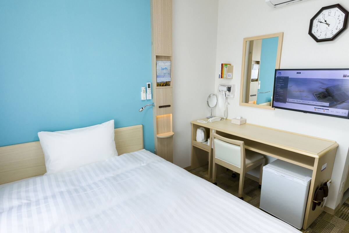 A hotel room with a bed, a desk with a chair, a small refrigerator, a mirror, a clock, and a TV on the wall.
