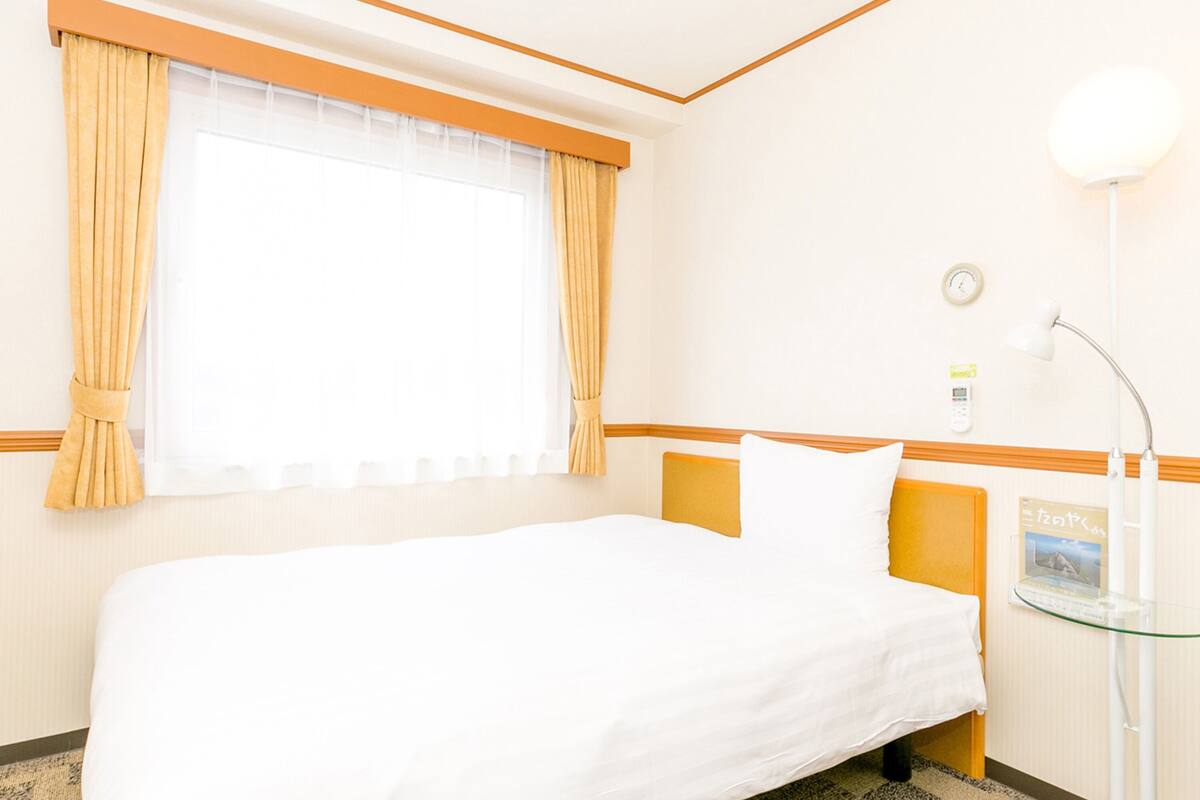 Una habitación de hotel con cama, una mesita de noche con lámpara, una ventana con cortinas y un reloj de pared.
