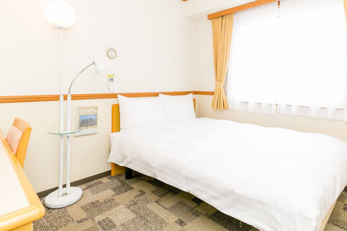 Habitación de hotel con una cama individual, una mesita de noche, una lámpara, un reloj y una ventana con cortinas.