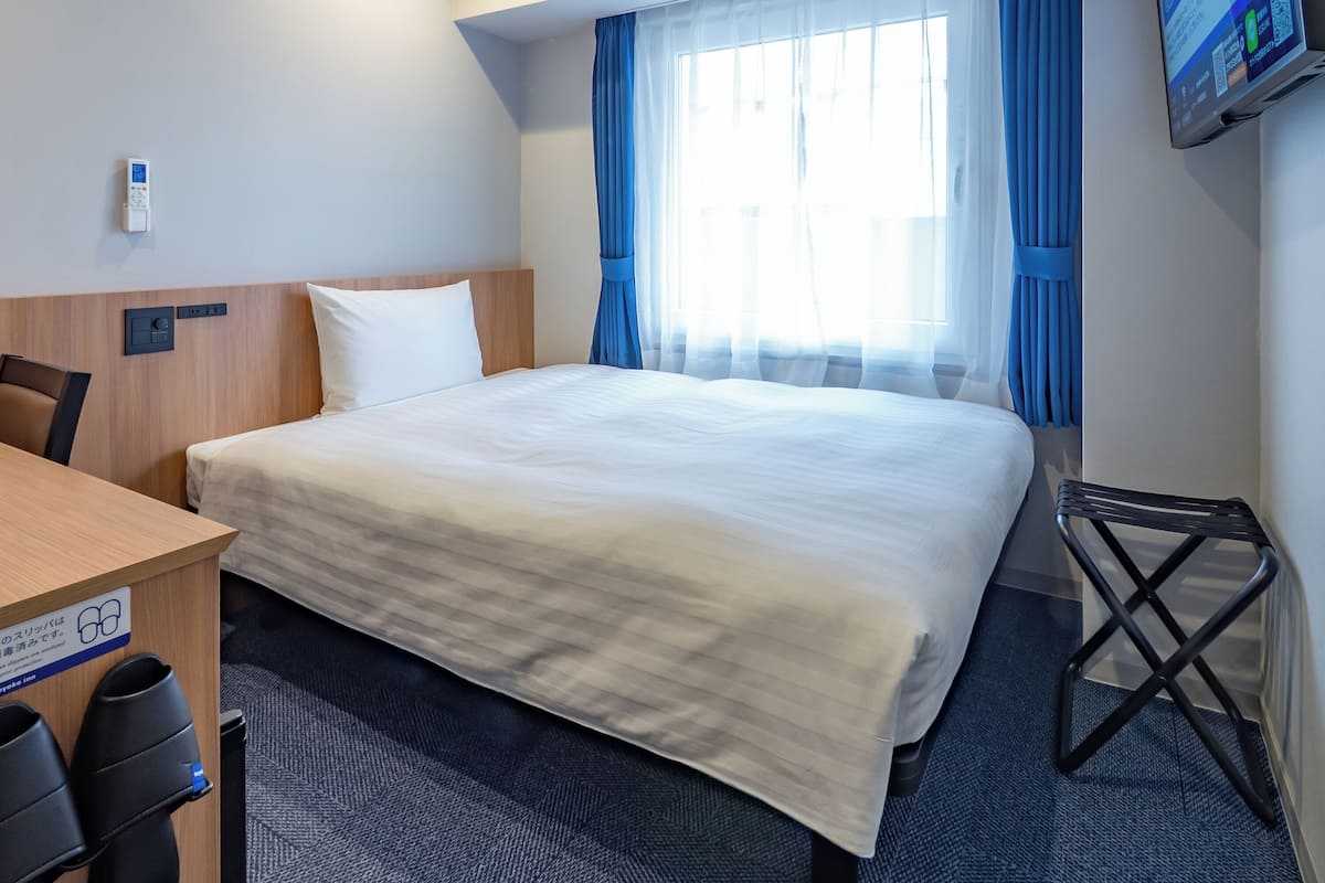 A hotel room with a bed, a desk, a chair, a TV mounted on the wall, and a window with curtains.