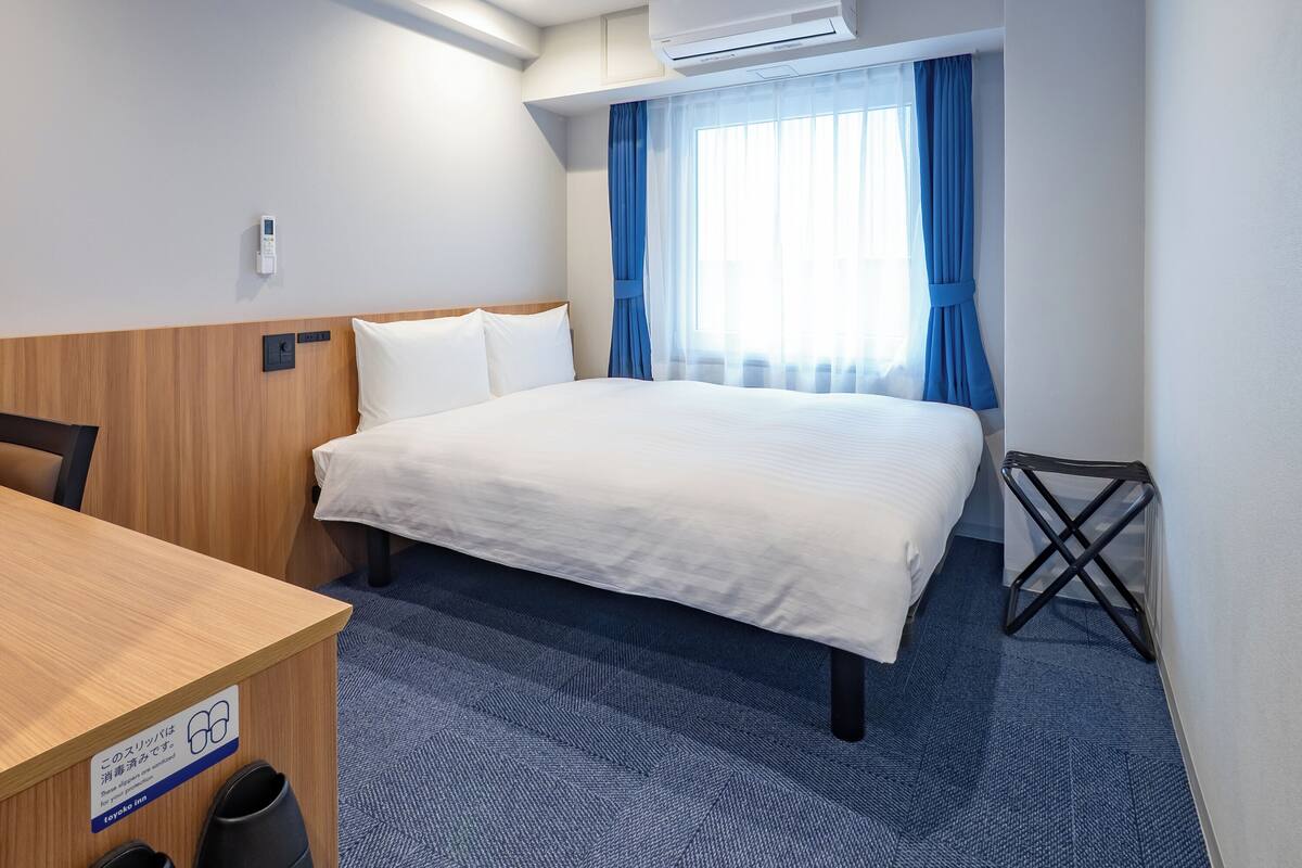 A hotel room with a bed, a desk, a chair, an air conditioning unit, and a window with curtains.