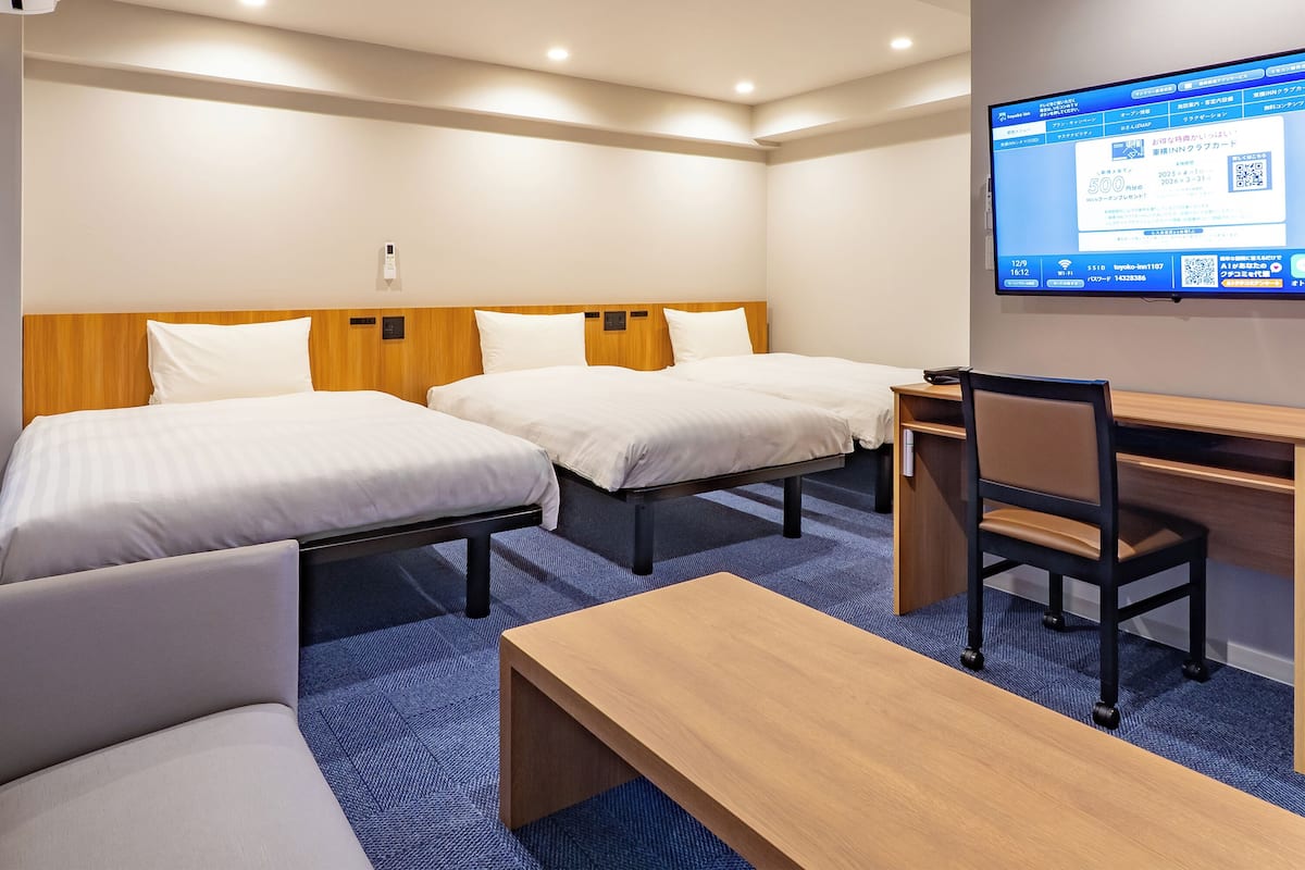 A hotel room with two beds, a desk, a chair, and a TV displaying a booking interface.