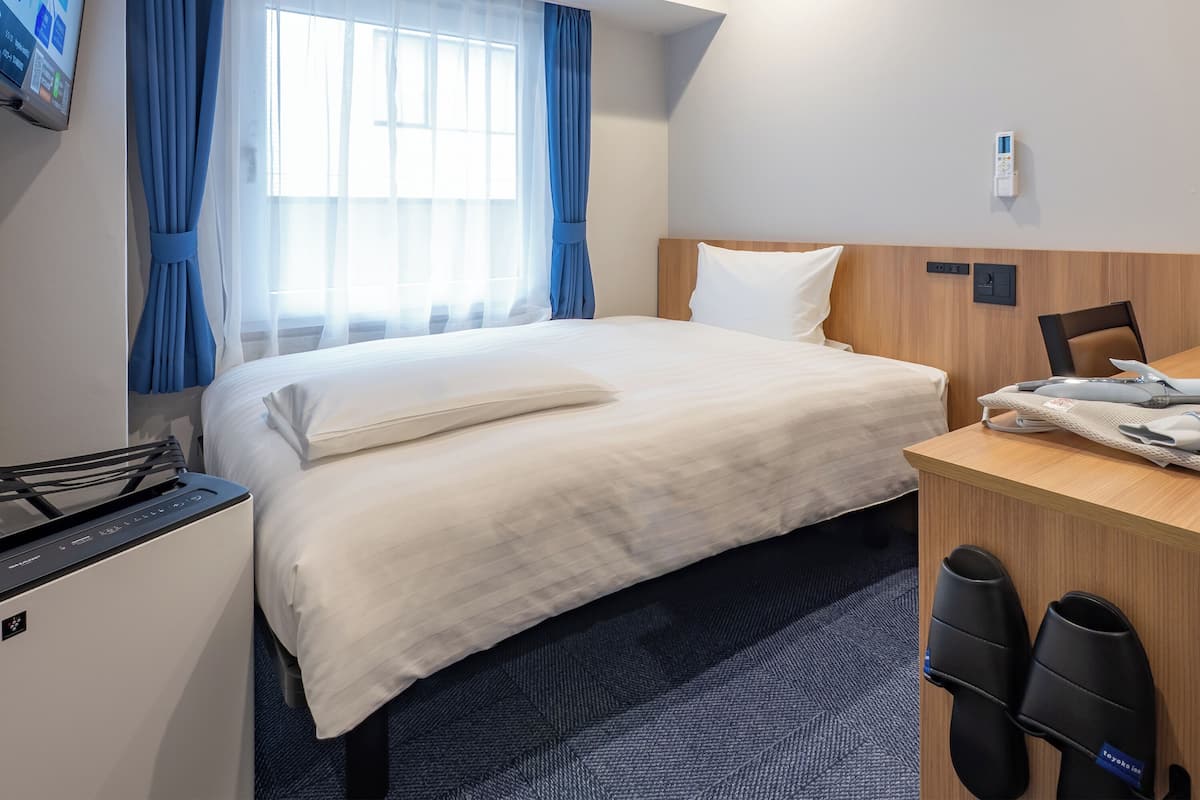 A hotel room with a bed, a desk, a chair, a small table, a heater, and a window with curtains.