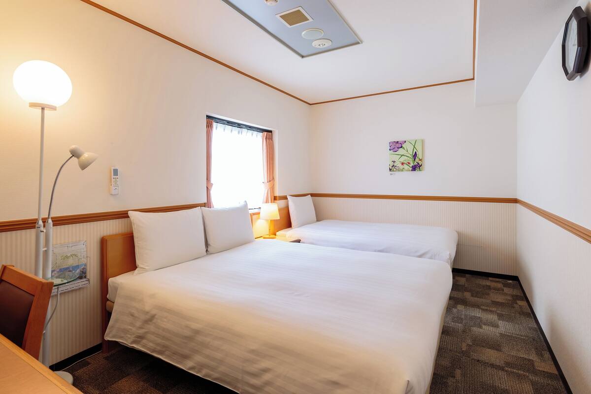 A hotel room with two beds, a desk, a lamp, a window with curtains, a wall clock, and a small painting.