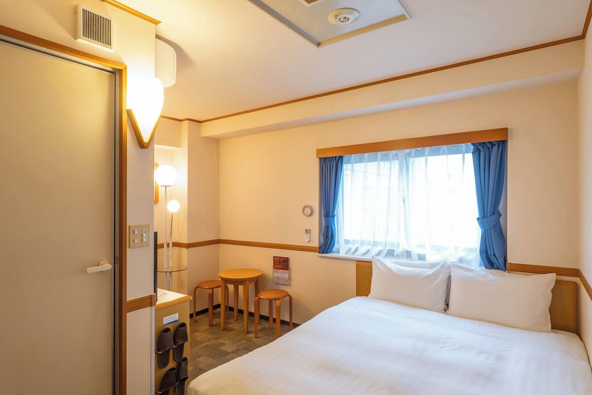 Hotelkamer met een groot bed, een klein tafeltje met twee stoelen, een spiegel en een raam met gordijnen.