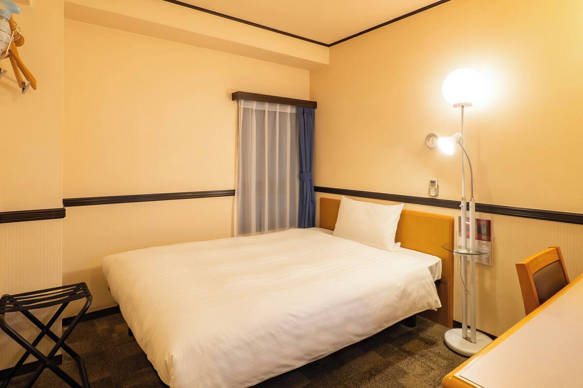 Een hotelkamer met een bed, bureau, stoel, lamp en een raam met gordijnen.