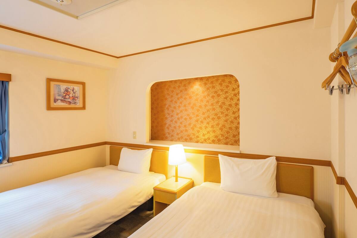 Een hotelkamer met twee enkele bedden, een nachtkastje met lamp, een ingelijst schilderij aan de muur en een raam met gordijnen.