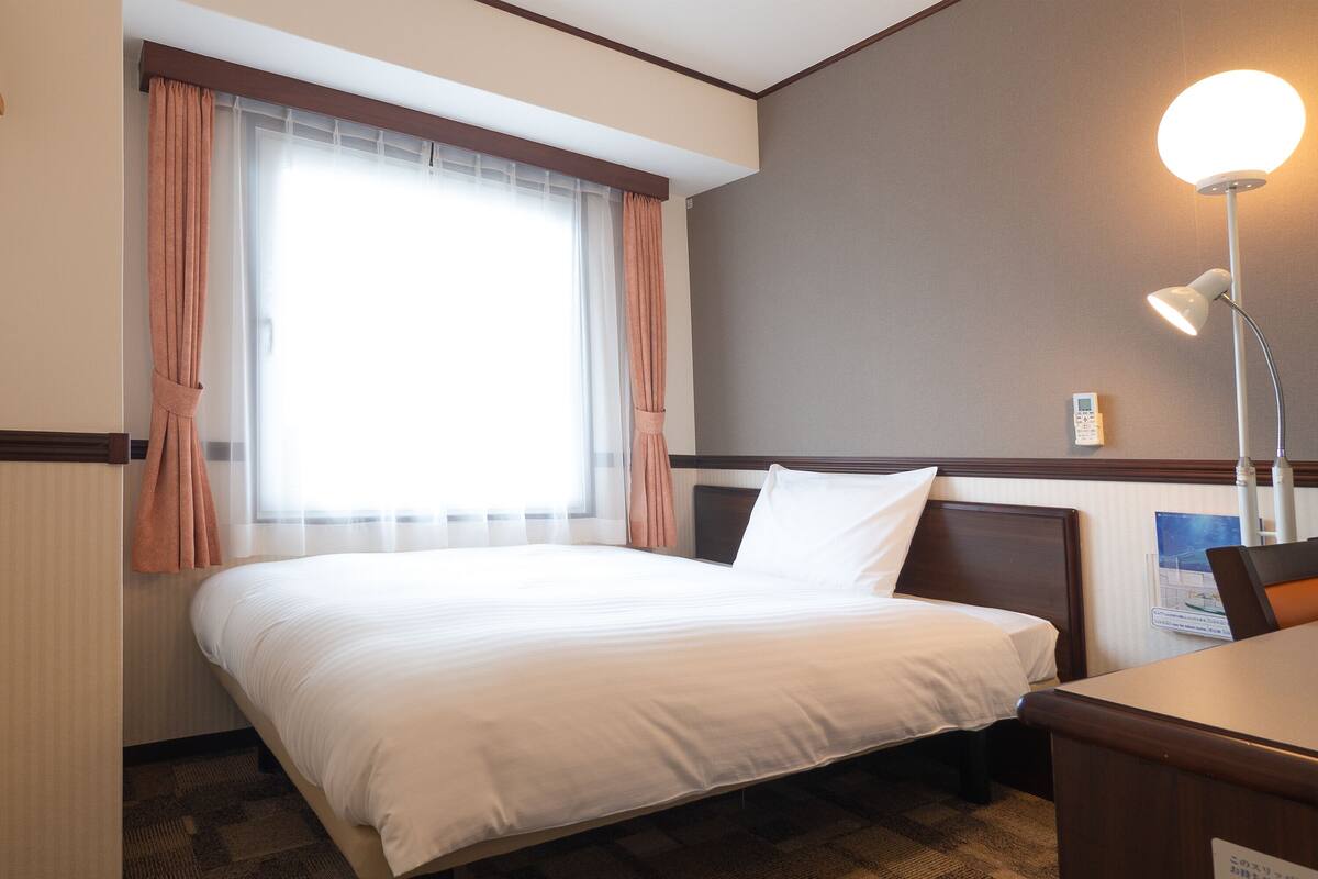 Ein Hotelzimmer mit Bett, Schreibtisch, Lampe und einem Fenster mit Vorhängen.