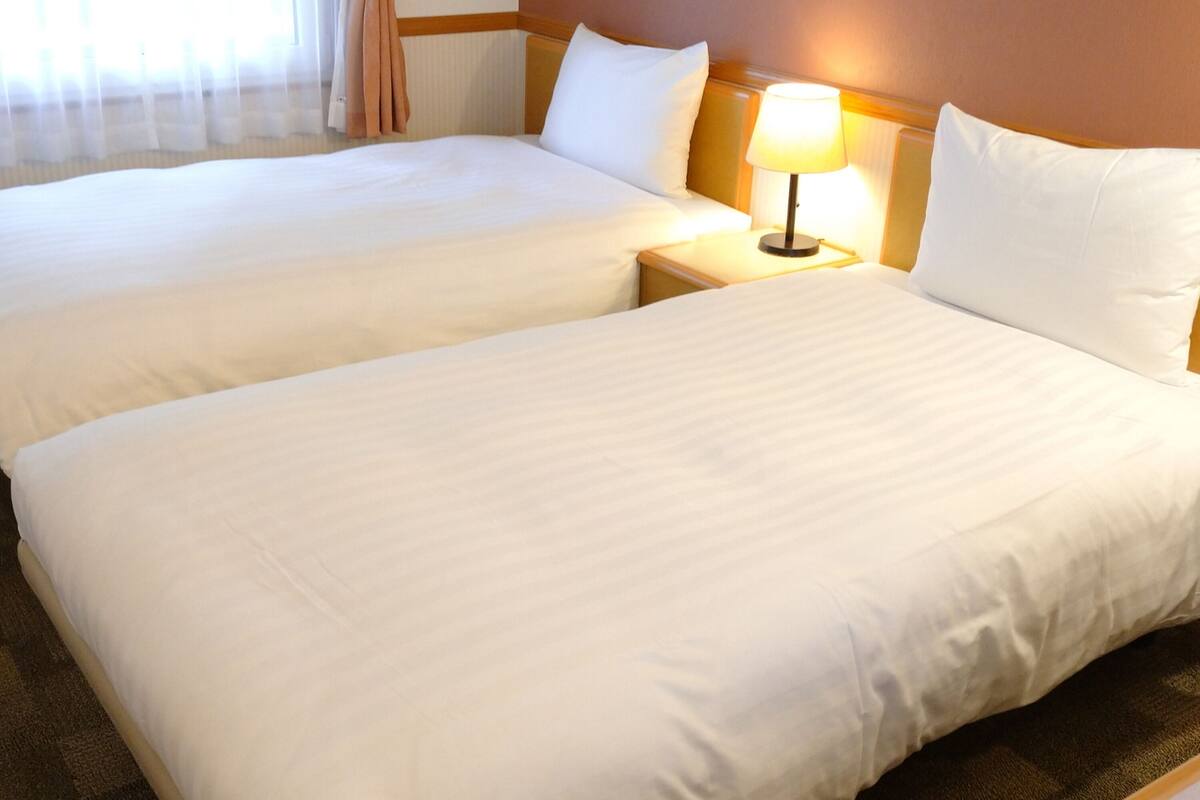 Two single beds with white linens, a bedside lamp, and a window with curtains.