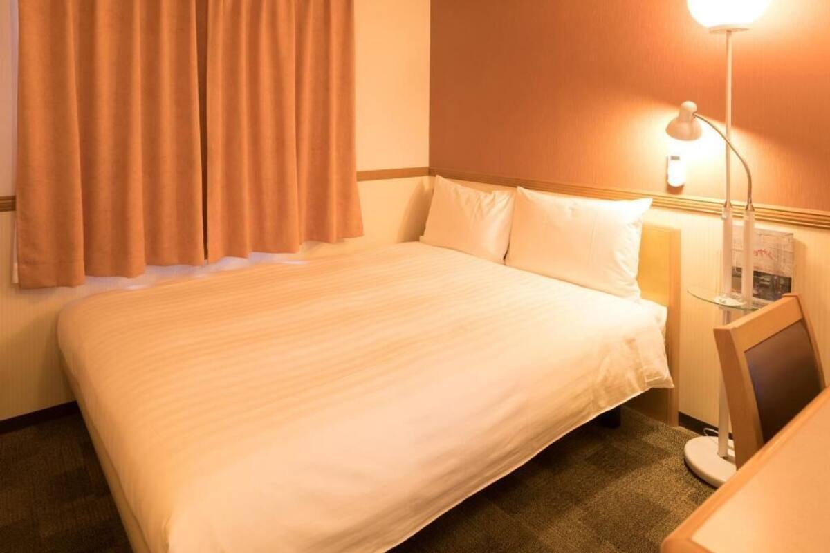 Een hotelkamer met een bed, gordijnen, een bureau met een lamp en een aan de muur bevestigde televisie.