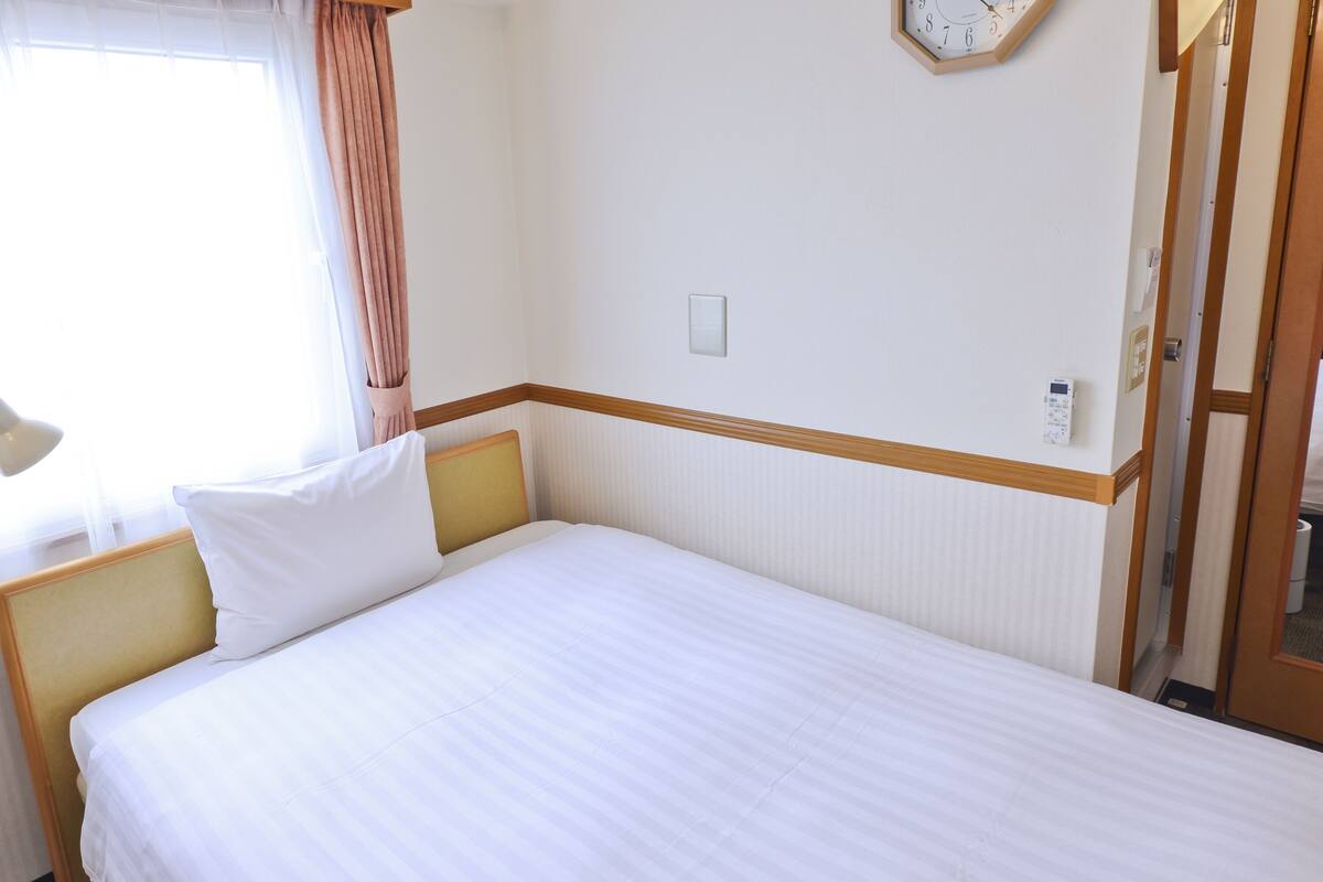 Hotelkamer met een eenpersoonsbed, nachtkastje, lamp, klok aan de muur en een raam met gordijnen.