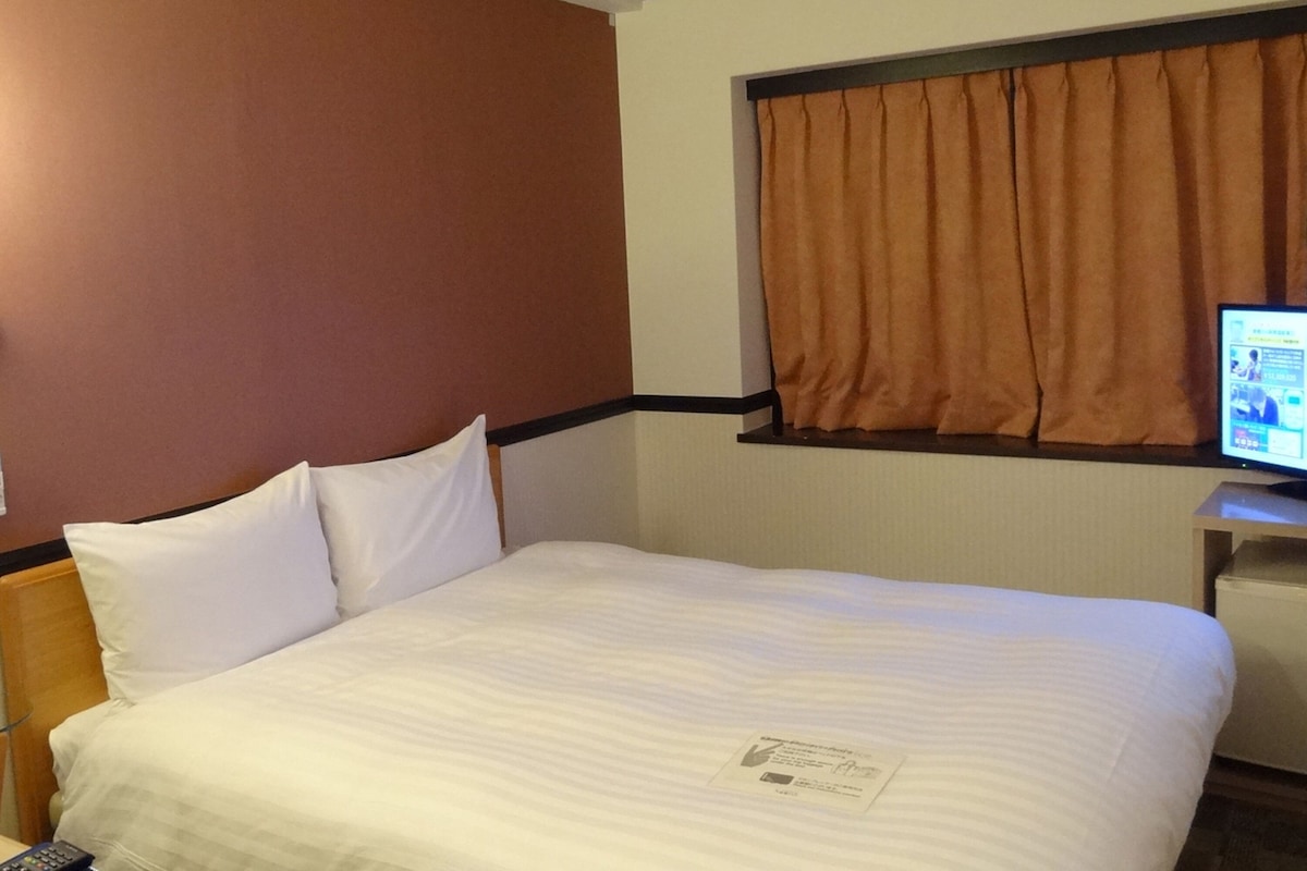 Een hotelkamer met een bed, kussens, een afstandsbediening, een televisie op een standaard en een raam met gordijnen.