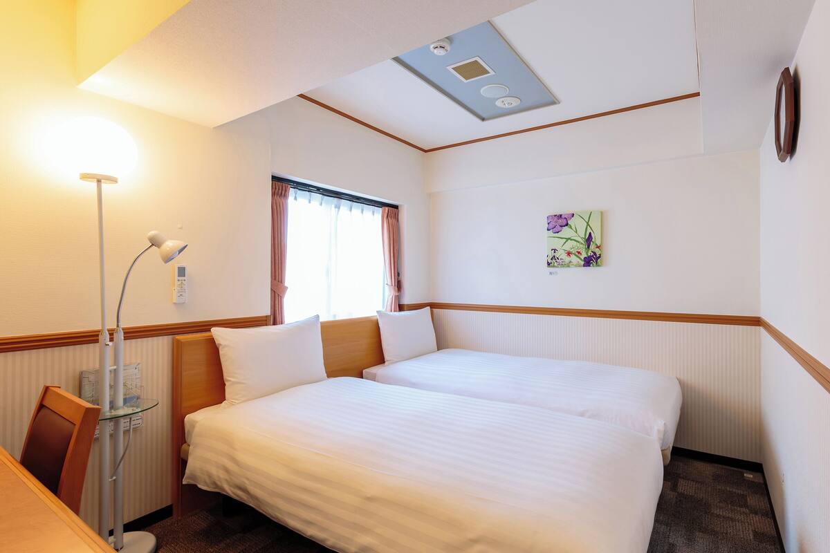 A hotel room with a bed, bedside table, lamp, window, and a painting on the wall.