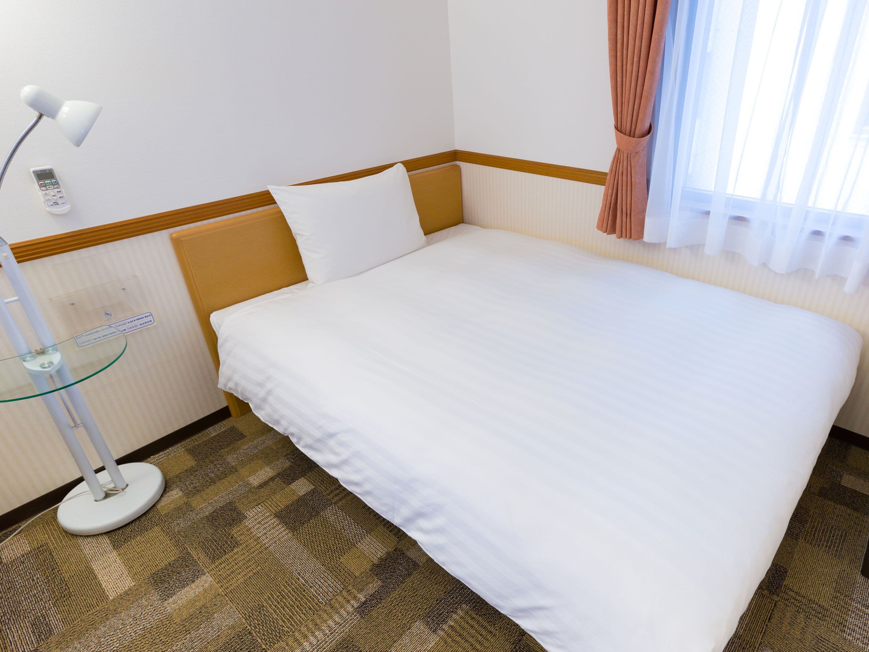 A hotel room with a bed, a bedside table with a lamp, a remote control, and a window with curtains.