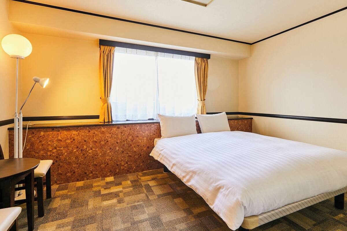 A hotel room with a bed, a desk, a lamp, and a window with curtains.