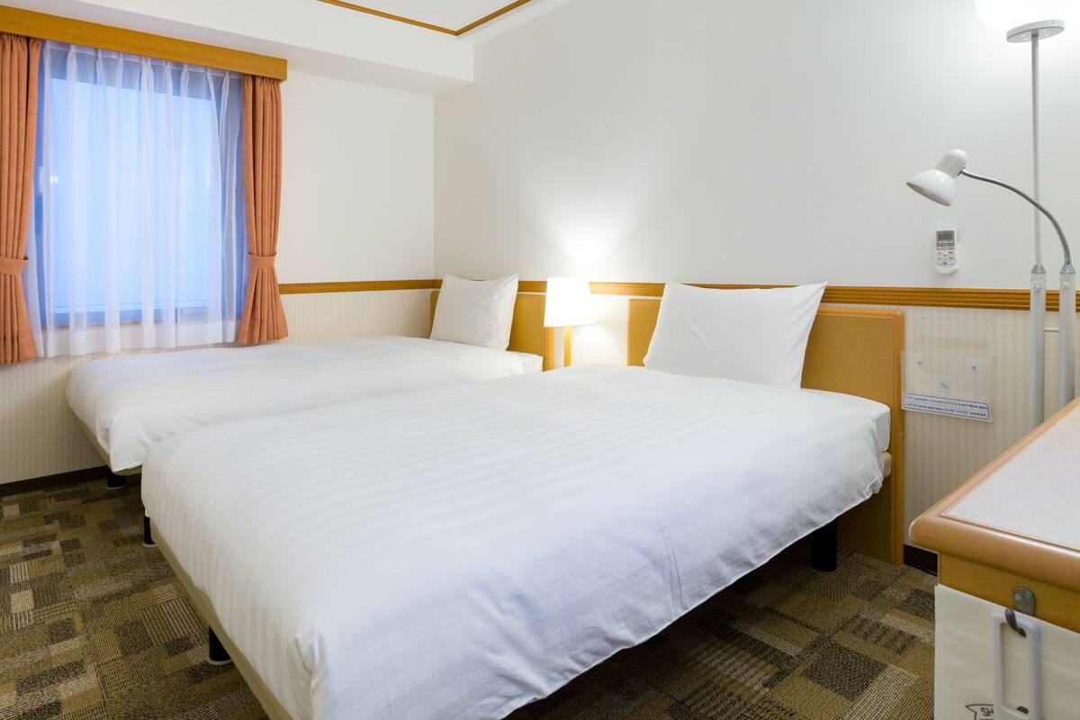 A hotel room with two single beds, a bedside table with a lamp, a window with sheer curtains, and a wall-mounted control panel.
