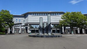 A modern building with a central fountain, large windows, and a parking lot. - Toyoko Inn Sendai Higashi 2 (Sendai)