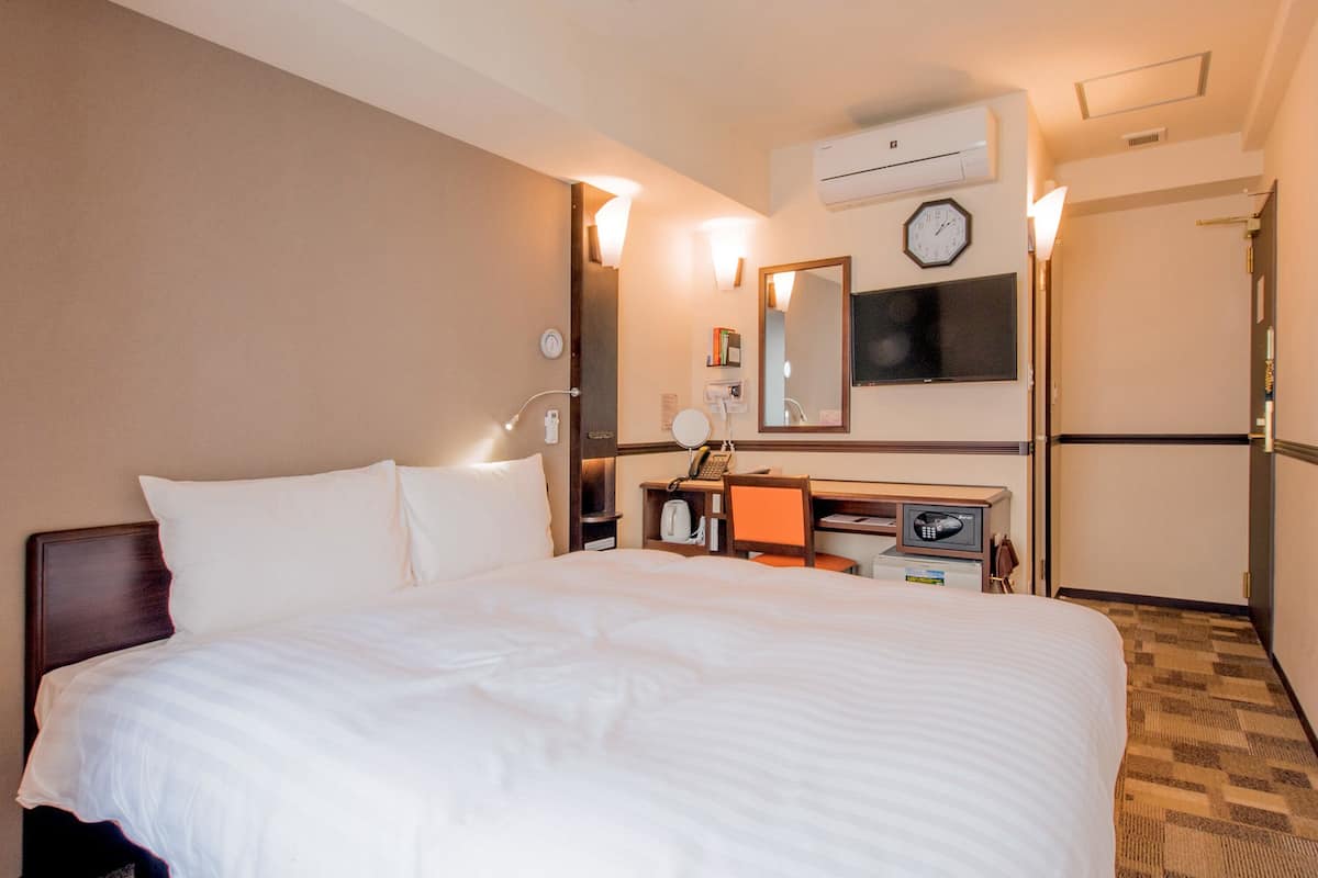 A hotel room with a large bed, a desk with a computer, a mirror, a clock, and an air conditioning unit.