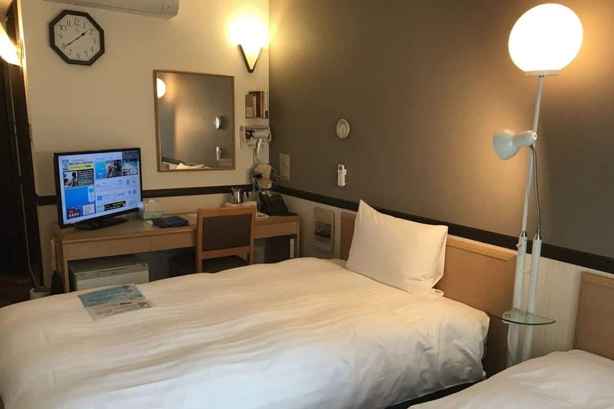 A hotel room with two beds, a desk with a computer, a chair, a desk lamp, and a clock on the wall.