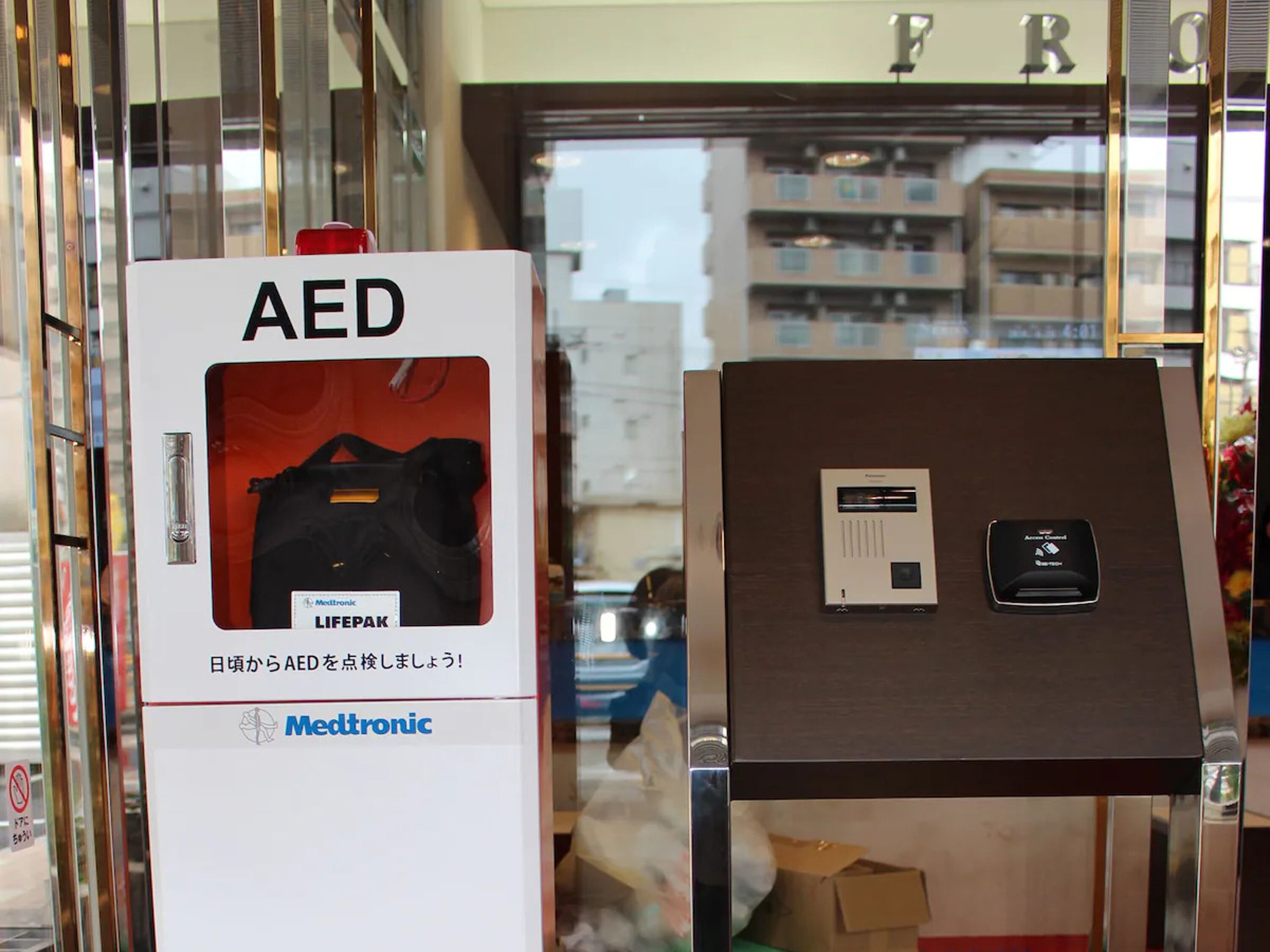 A display of AED devices in a store with a glass door and a view of buildings outside.