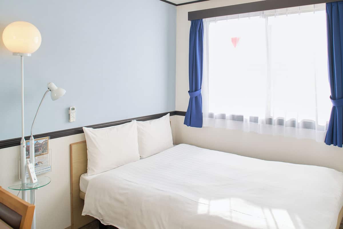 Una cama bien hecha con sábanas blancas en una habitación con pared azul, una mesita de noche con lámpara y una ventana con cortinas azules.