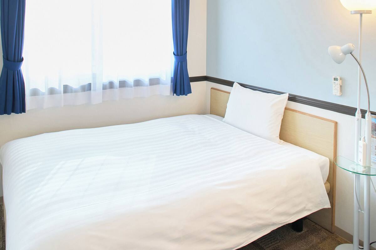 Habitación de hotel con una cama bien hecha, una mesita de noche con lámpara y una ventana con cortinas.