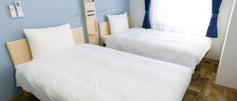 Quarto de hotel com duas camas, cabeceira de madeira, mesa de cabeceira, luminária e janela com cortinas.