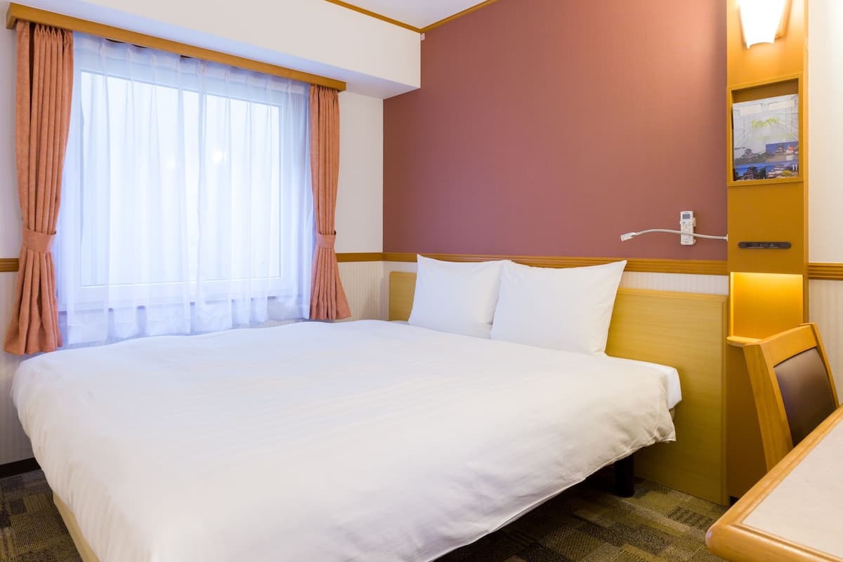 A hotel room with a large bed, a desk, a window with curtains, and a wall-mounted light.