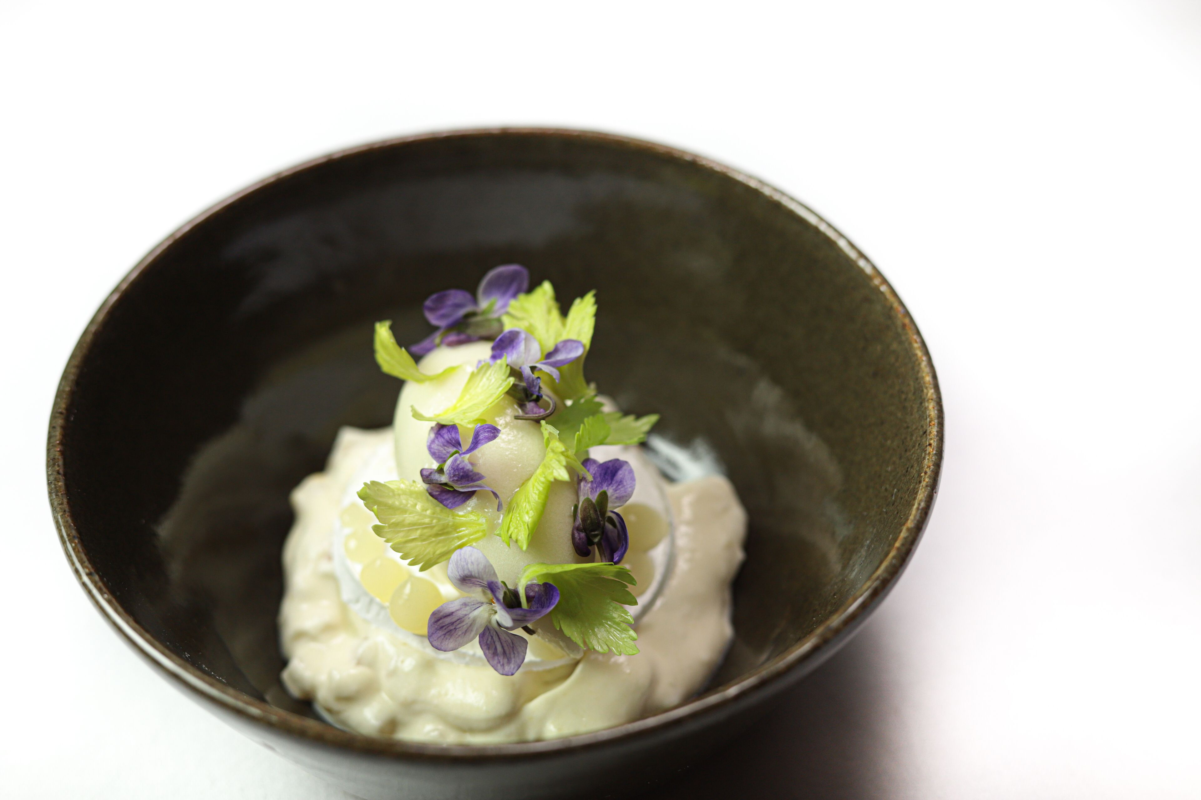 A bowl of creamy dessert with purple flowers and green leaves on a dark ceramic plate.