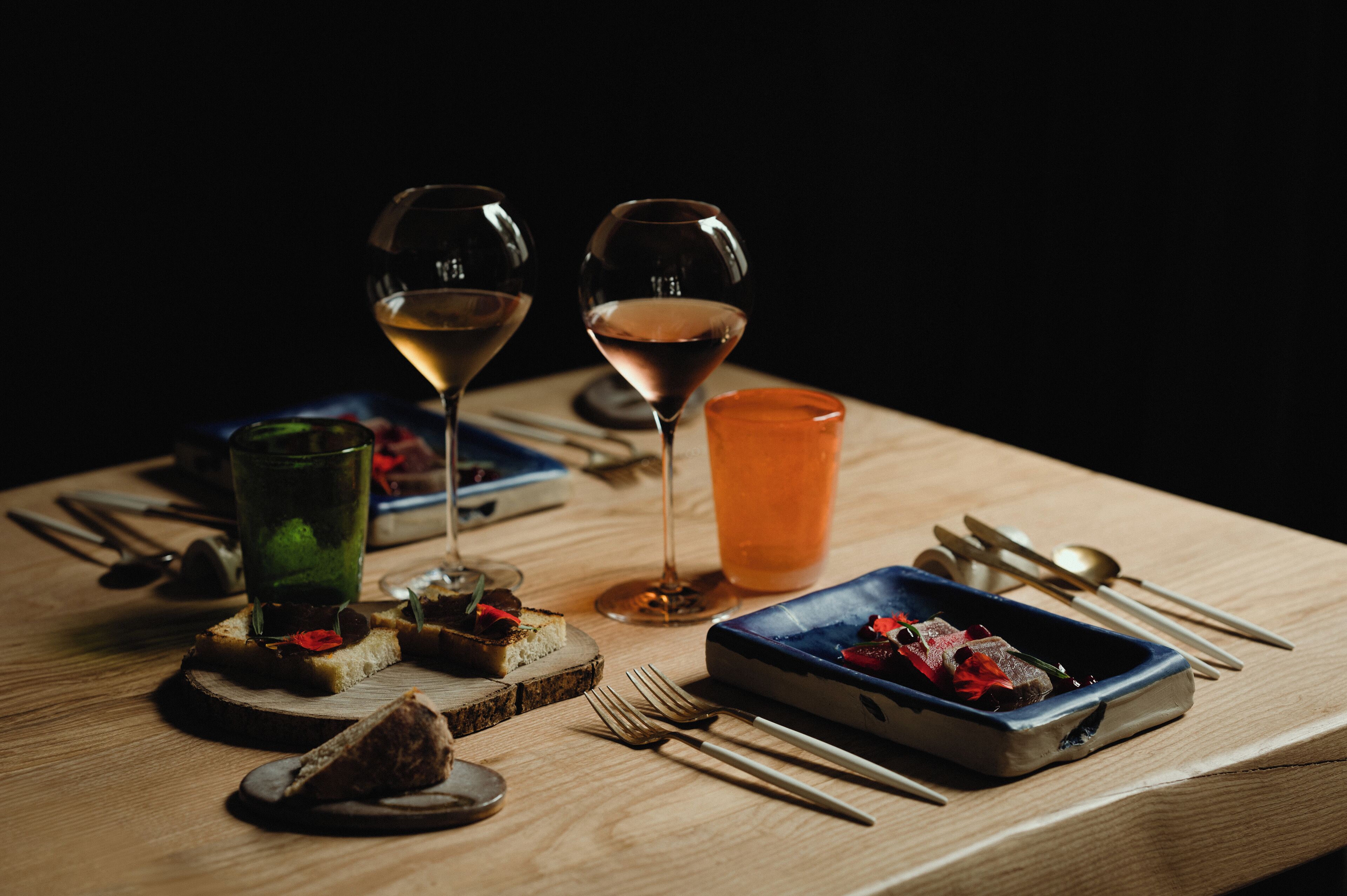 A wooden table with a blue dish containing food, two wine glasses, a green glass, and a red glass.