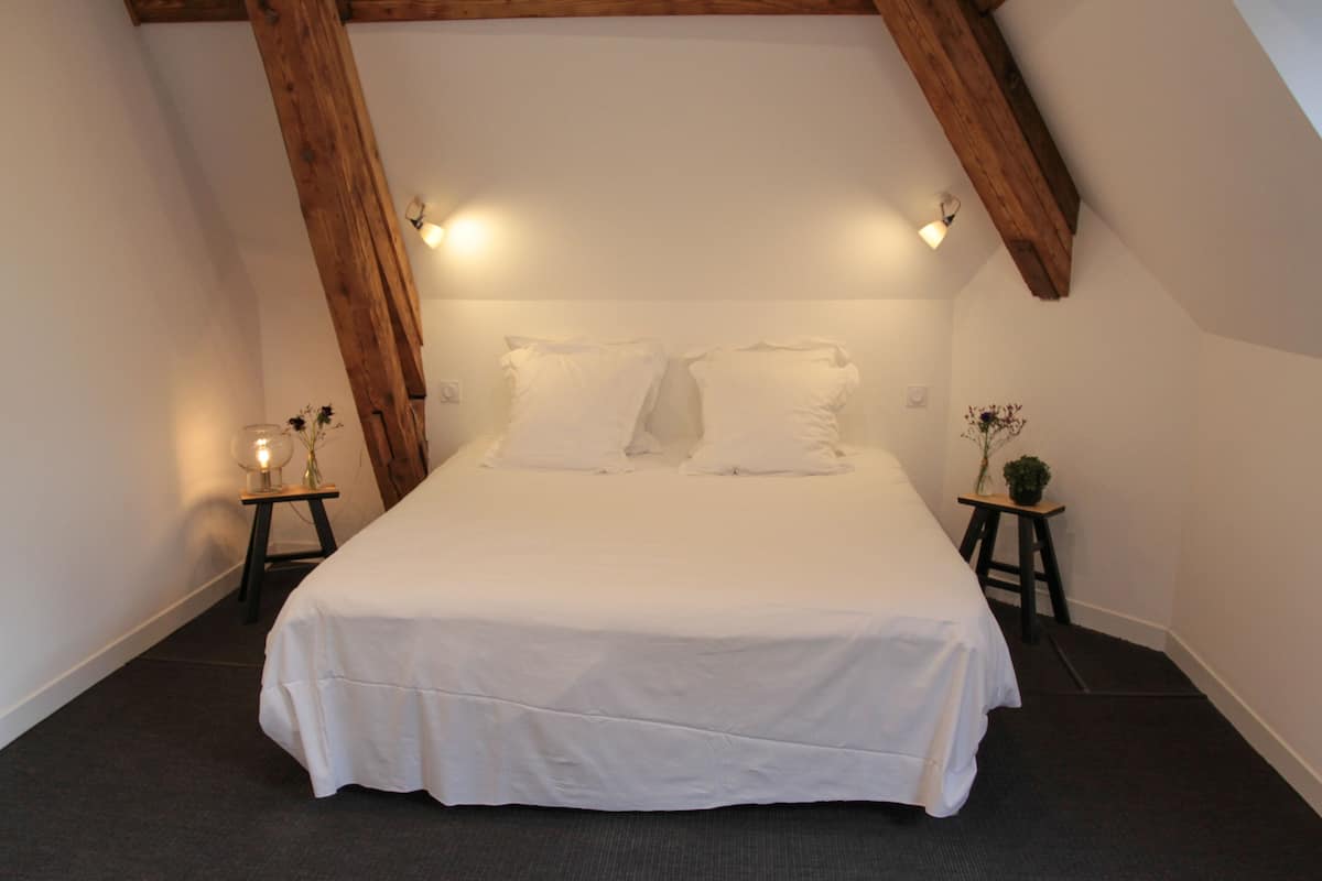 Un dormitorio con una cama grande, vigas de madera, dos mesitas de noche con lámparas y una planta pequeña.