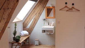 Ein kleines, gemütliches Schlafzimmer unter dem Dach mit geneigter Decke, Holzbalken, einem Oberlicht, einer Holzbank, einem kleinen Tisch, einer Topfpflanze, einem weißen Waschbecken, einem Spiegel und zwei Holzaufhängern an der Wand.