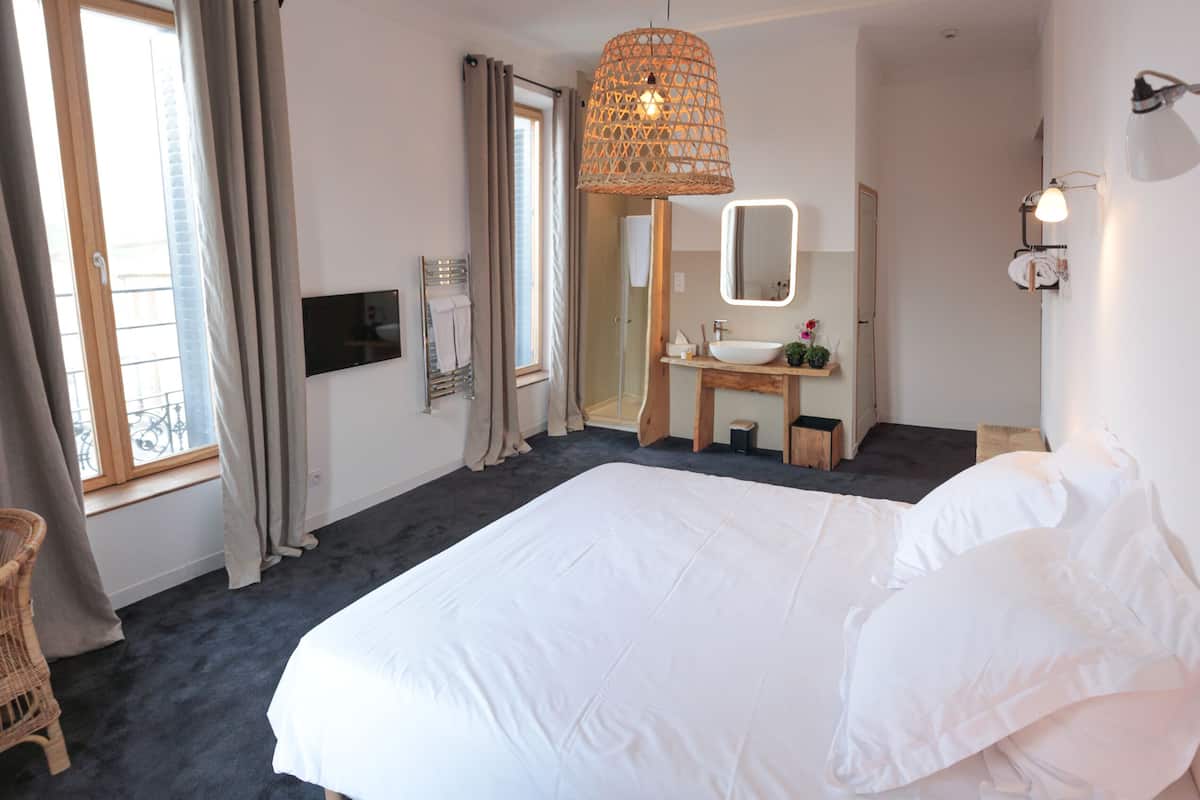 Habitación de hotel con una cama grande, una silla de mimbre, un baño con lavamanos y espejo, y una ventana con cortinas.