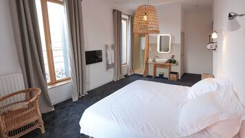 A hotel room with a large bed, a wicker chair, a bathroom with a sink and mirror, and a window with curtains.