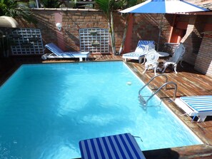 A rectangular swimming pool with a wooden deck, surrounded by lounge chairs and a small table. - Pousada Brilho Da Serra (Tiradentes)