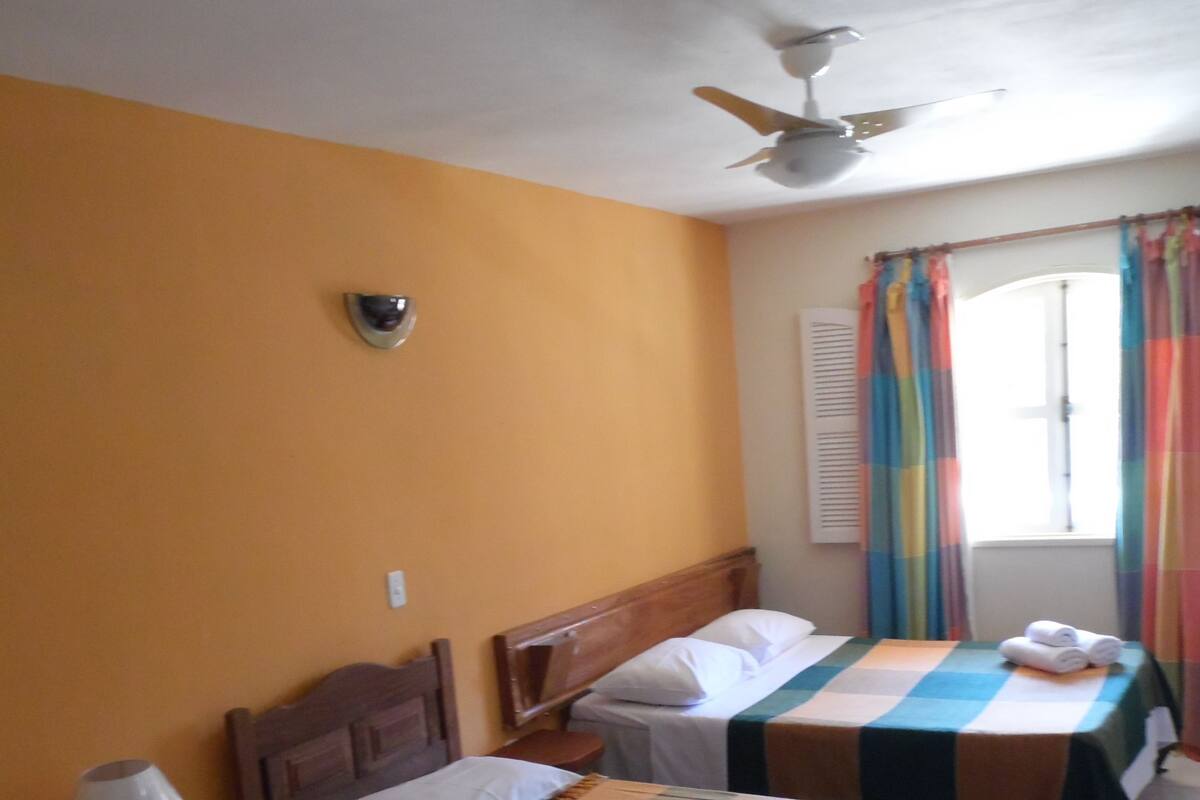 A room with two single beds, a ceiling fan, a window with colorful curtains, and a small table with a lamp.