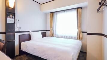 A hotel room with a bed, a desk, a chair, a window with curtains, and a wall-mounted light fixture.