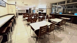 Une cafétéria spacieuse, dotée de nombreuses tables et chaises, d’un comptoir avec un ordinateur et d’une grande fenêtre offrant une vue sur l’extérieur.
