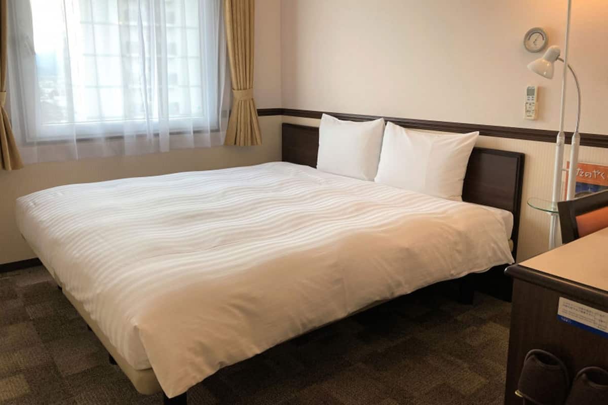 A hotel room with a large bed, a bedside table, a lamp, a clock, and a window with curtains.