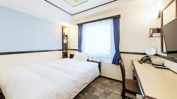 Una habitación de hotel con una cama, un escritorio con teléfono y espejo, una ventana con cortinas y una repisa con un cartel.