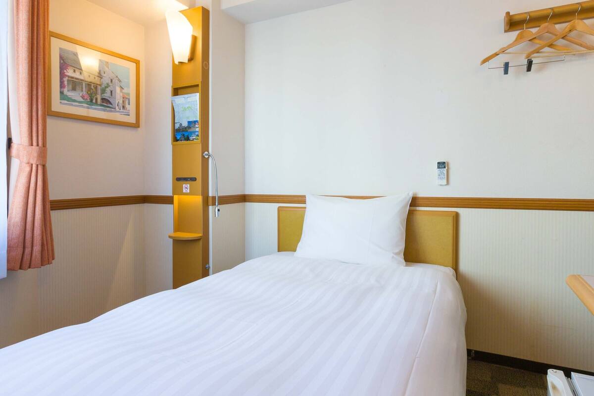 A hotel room with a bed, a wooden headboard, a wall-mounted light, a framed picture, and a wooden hanger rack.