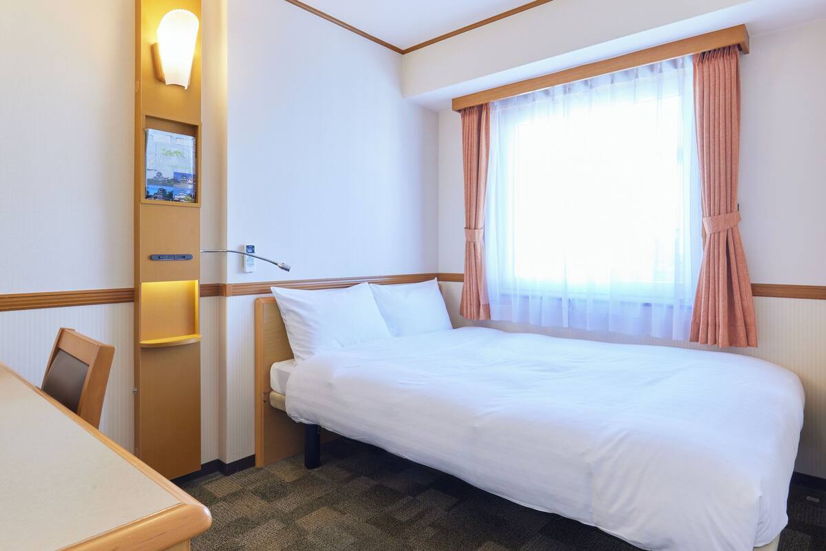 A hotel room with a bed, a desk, a chair, a wall-mounted lamp, and a window with curtains.